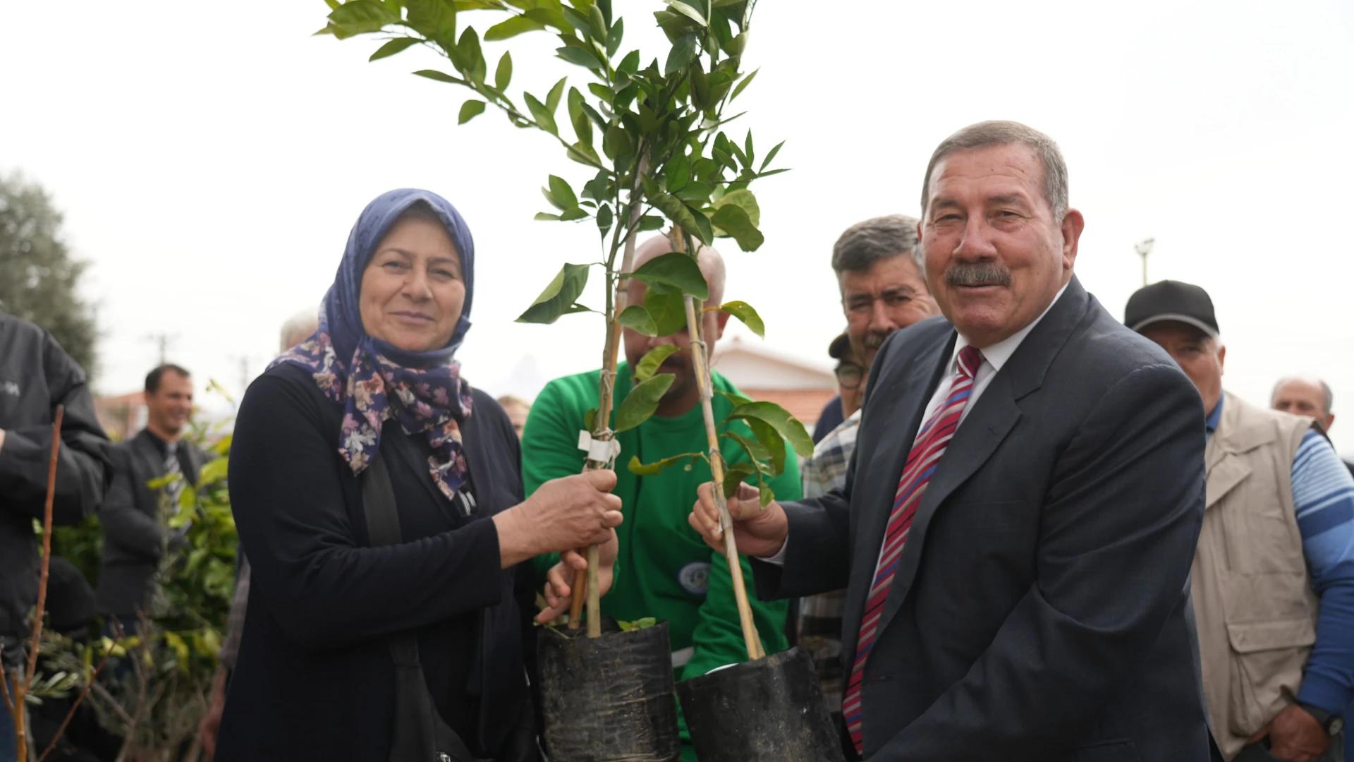 Bir kadın ve takım elbiseli bir erkek, kalabalık önünde saksılı fidanları birlikte tutuyor.