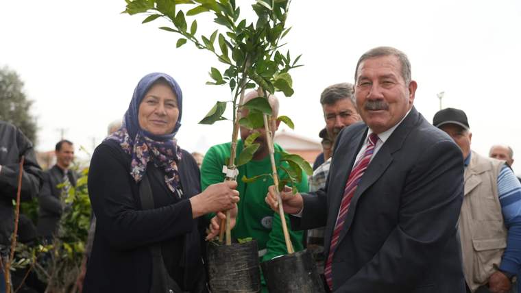 Bir kadın ve takım elbiseli bir erkek, kalabalık önünde saksılı fidanları birlikte tutuyor.