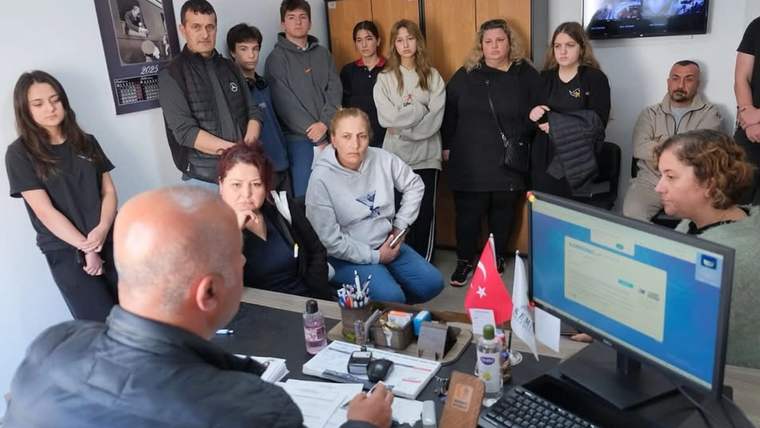 Masa başında oturan bir kişi, bilgisayar ekranı önünde; çevresinde ayakta duran ve oturan kişiler bulunuyor.