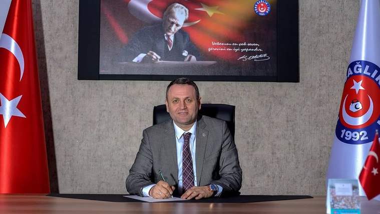 Masada oturan takım elbiseli bir kişi, arkasındaki Türk bayrağı temalı pano ve iki bayrak önünde duruyor.