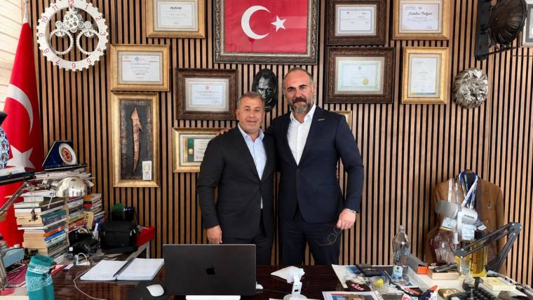Çerçeveli belgeler ve Türk bayrağı bulunan bir odada, masanın arkasında yan yana duran iki kişi