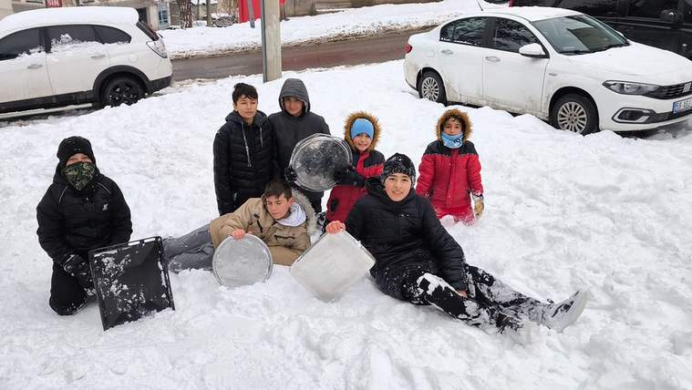 Karla kaplı alanda oturan çocuklar, tepsilerle eğleniyorlar.