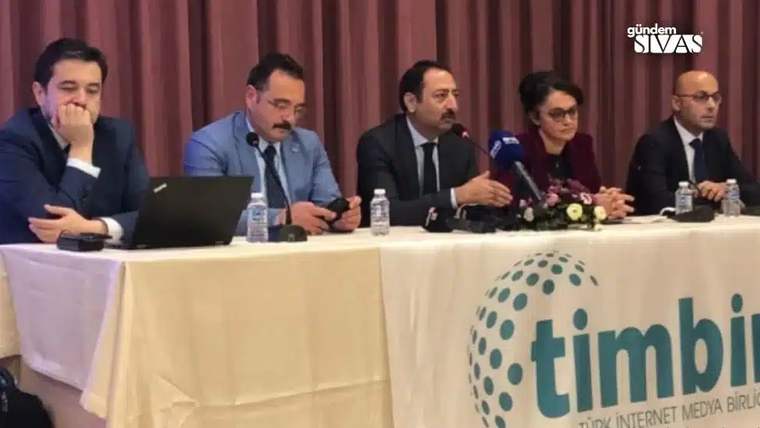 Masa başında oturan beş kişi, mikrofonlar ve su şişeleriyle bir panelde konuşuyor; önde logolu afiş bulunuyor.