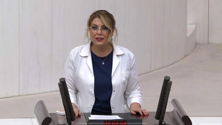 Beyaz önlüklü bir kadın, meclis kürsüsünde iki mikrofonun önünde konuşuyor.