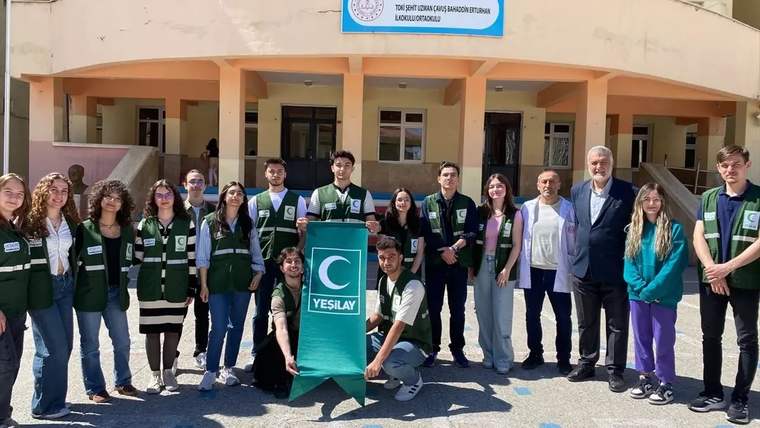 Okul binası önünde Yeşilay yazılı pankartı tutan, yeşil yelekli ve sivil giyimli bir grup duruyor.