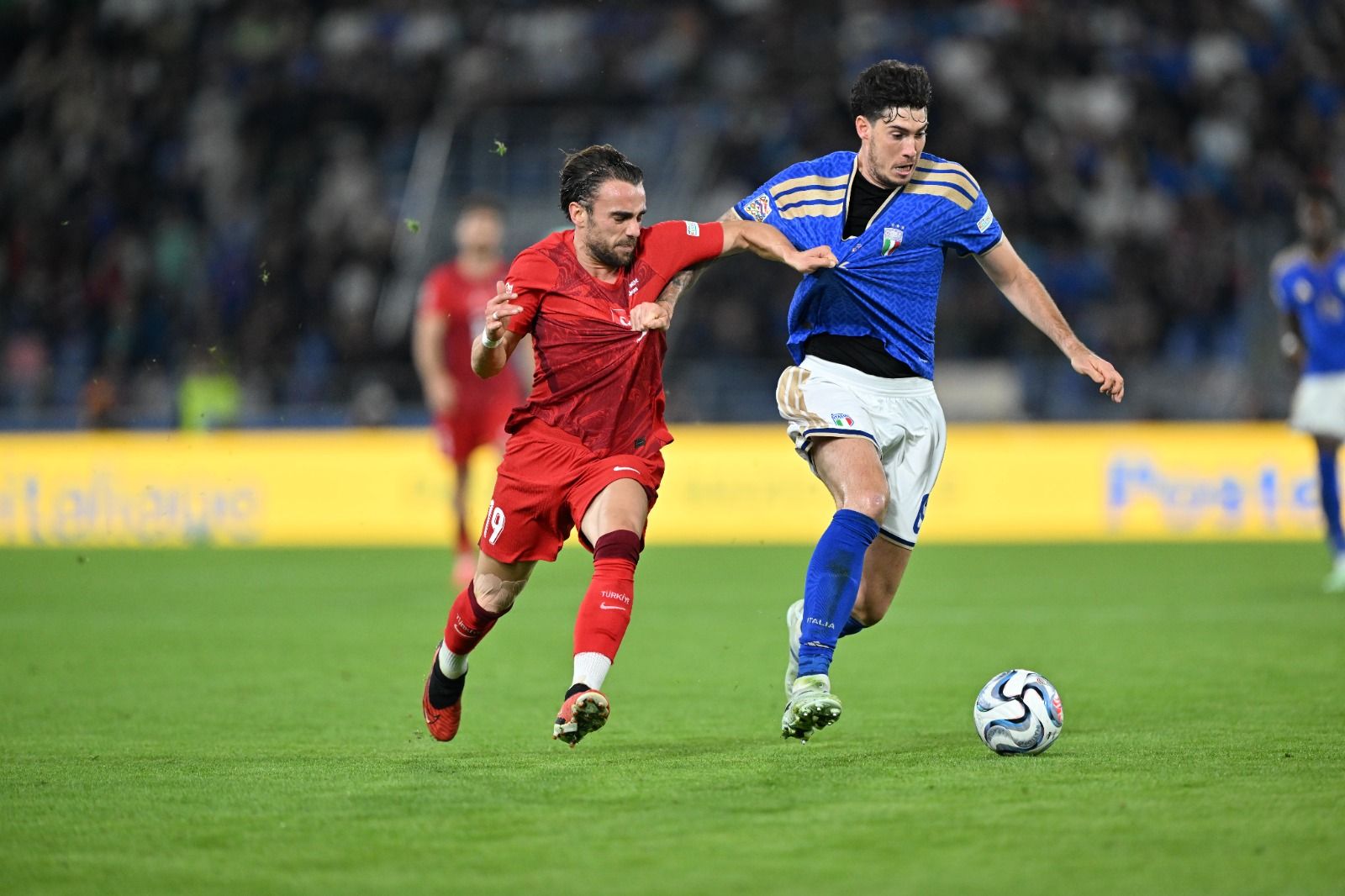 Kırmızı formalı oyuncu, mavi formalı rakibini geçmeye çalışırken futbol topu çim sahada ilerliyor.