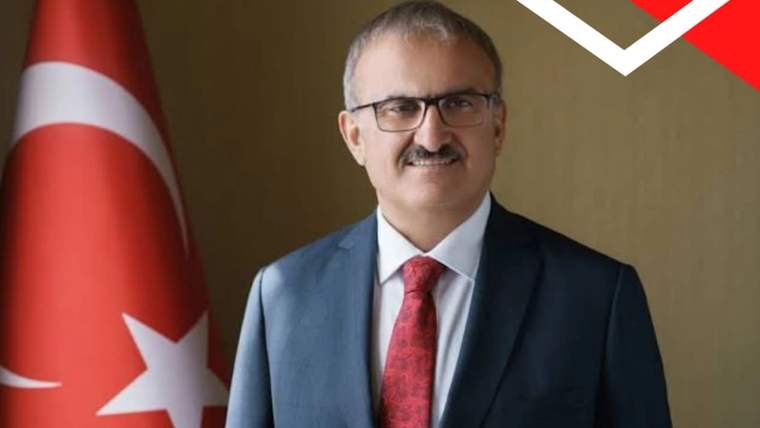 Bıyıklı ve gözlüklü bir erkek, takım elbise ve kırmızı kravatla Türk bayrağı yanında duruyor.