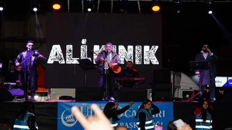 Gece sahnesinde mikrofon başında gitar çalan bir kişi, yanında müzisyenler ve arkada “ALİ KINIK” yazısı görülüyor.
