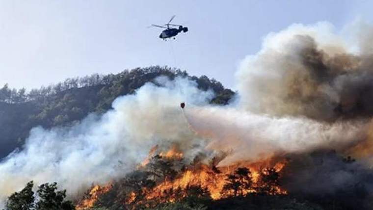 Ormanlık alandaki alevlerin üzerinde uçan helikopter, yangına su boşaltırken yoğun duman yükseliyor.