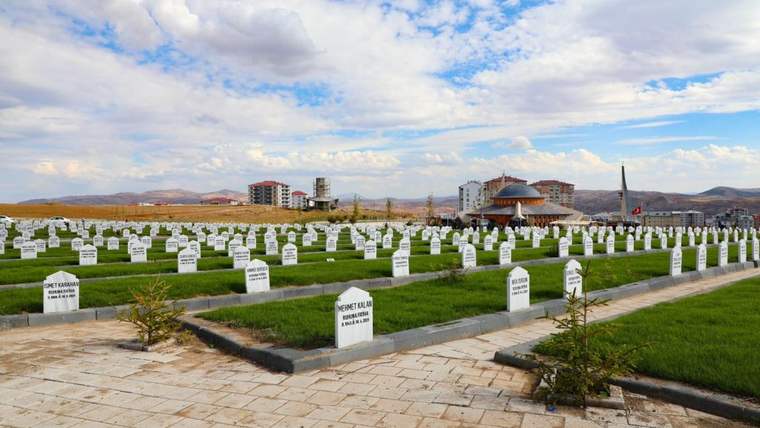 Beyaz mezar taşlarının sıralandığı bir mezarlık, arka planda binalar ve bulutlu bir gökyüzü görünmekte.