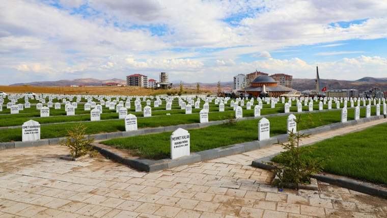 Beyaz mezar taşlarının sıralandığı bir mezarlık alanı, arka planda binalar ve bulutlu bir gökyüzü var.