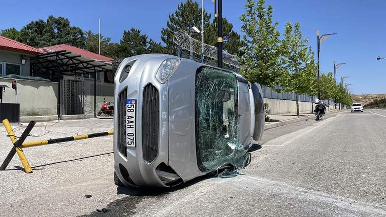 Yan yatmış gümüş renkli otomobilin camları kırılmış; yol kenarında araç parçaları ve cam kırıkları görülüyor.