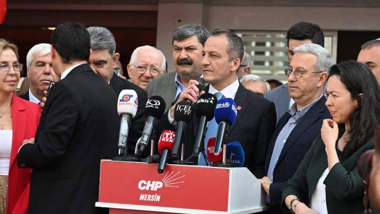 CHP Mersin yazılı kürsüde konuşan bir kişi, çok sayıda mikrofon ve çevresindeki kalabalıkla görülüyor.