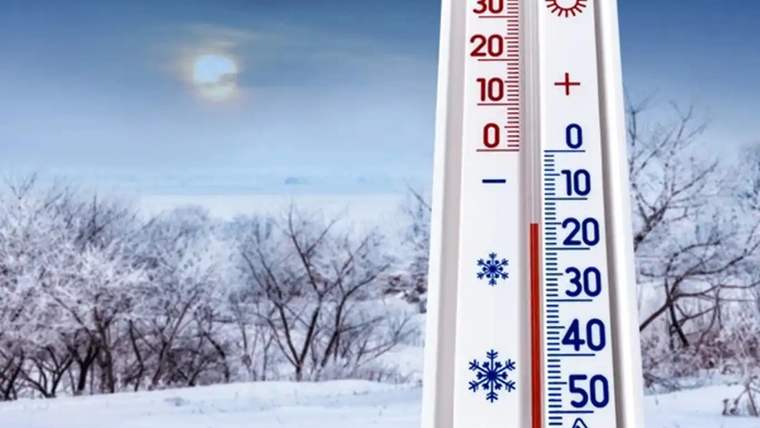 Bir termometre, eksi 25 dereceyi gösteriyor, arka planda karla kaplı ağaçlar ve güneşli bir gökyüzü var.