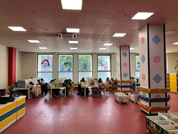 Çocuk ve yetişkinlerin masalarda oturduğu, kitaplıklarla çevrili geniş ve aydınlık kütüphane salonu.