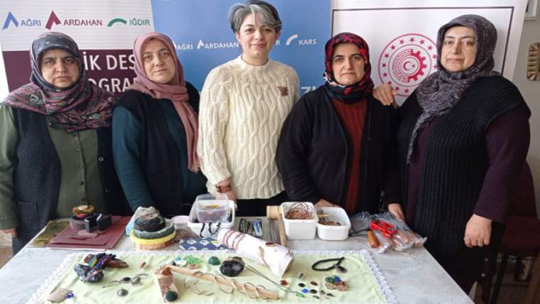 Beş kadın, masada sergilenen renkli el işi ürünleri ve yiyeceklerin arkasında yan yana duruyor.
