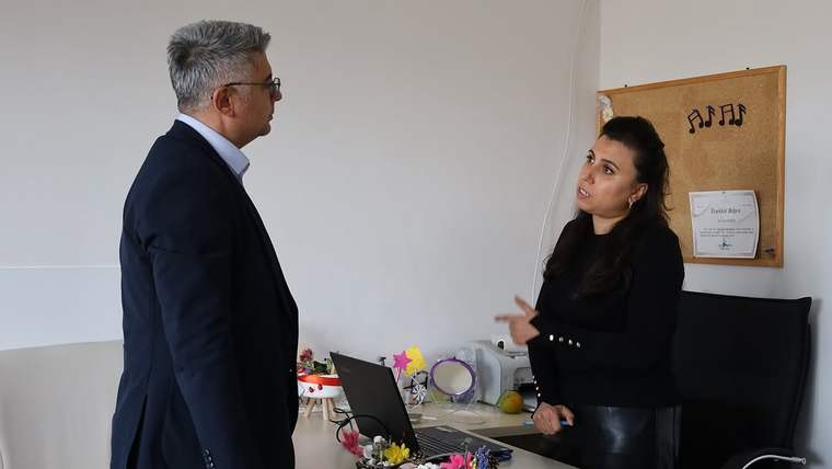 Bir ofiste ayakta duran iki yetişkin, çalışma masasının yanında konuşurken görülüyor.