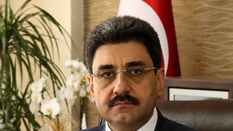 Gözlüklü ve bıyıklı bir kişi takım elbiseyle koltukta oturuyor; arka planda Türk bayrağı ve AK PARTİ yazısı görülüyor.