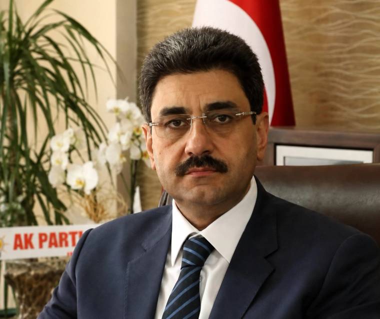 Gözlüklü ve bıyıklı bir kişi takım elbiseyle koltukta oturuyor; arka planda Türk bayrağı ve AK PARTİ yazısı görülüyor.