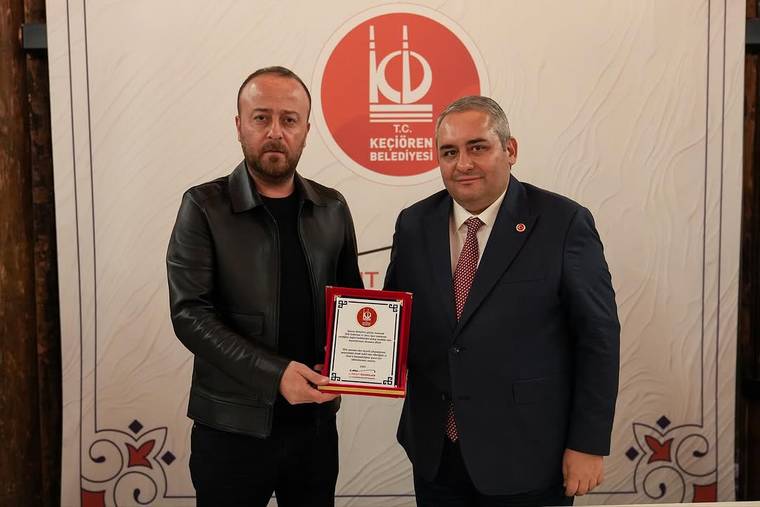 İki erkek, Keçiören Belediyesi logosunun önünde üzerinde yazılar bulunan bir plaketi birlikte tutuyor.