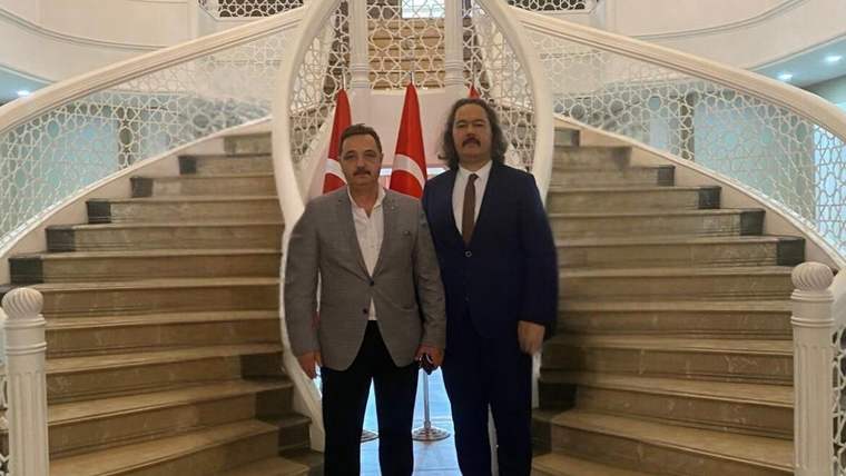 Süslemeli merdivenlerin önünde, arkalarında Türk bayrakları bulunan iki erkek yan yana duruyor.