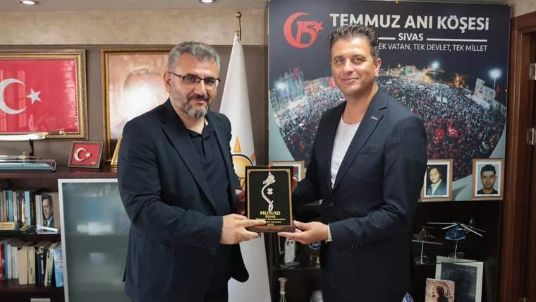Bir ofiste iki erkek, üzerinde MÜSİAD yazan plaketi tutuyor; arkalarında Türk bayrağı ve duvar görselleri bulunuyor.