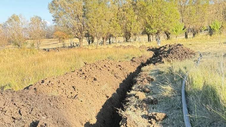 Toprağı kazılmış bir alan, yan tarafında su borusu görünmekte, arka planda ağaçlar var.