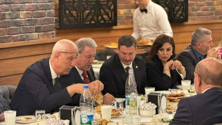 Bir grup insan, bir masada yemek yerken sohbet ediyor; arka planda garson hizmet veriyor.