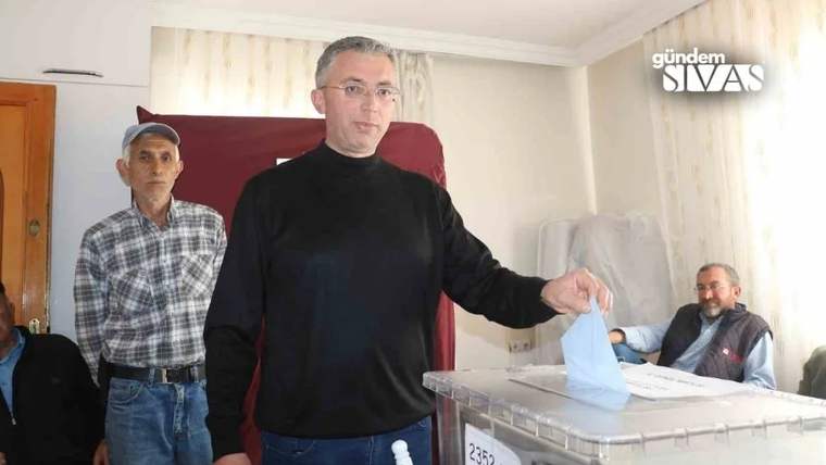 Bir kişi sandık başında oy pusulası tutarken, çevresinde üç kişi ve şeffaf oy sandığı görülüyor.