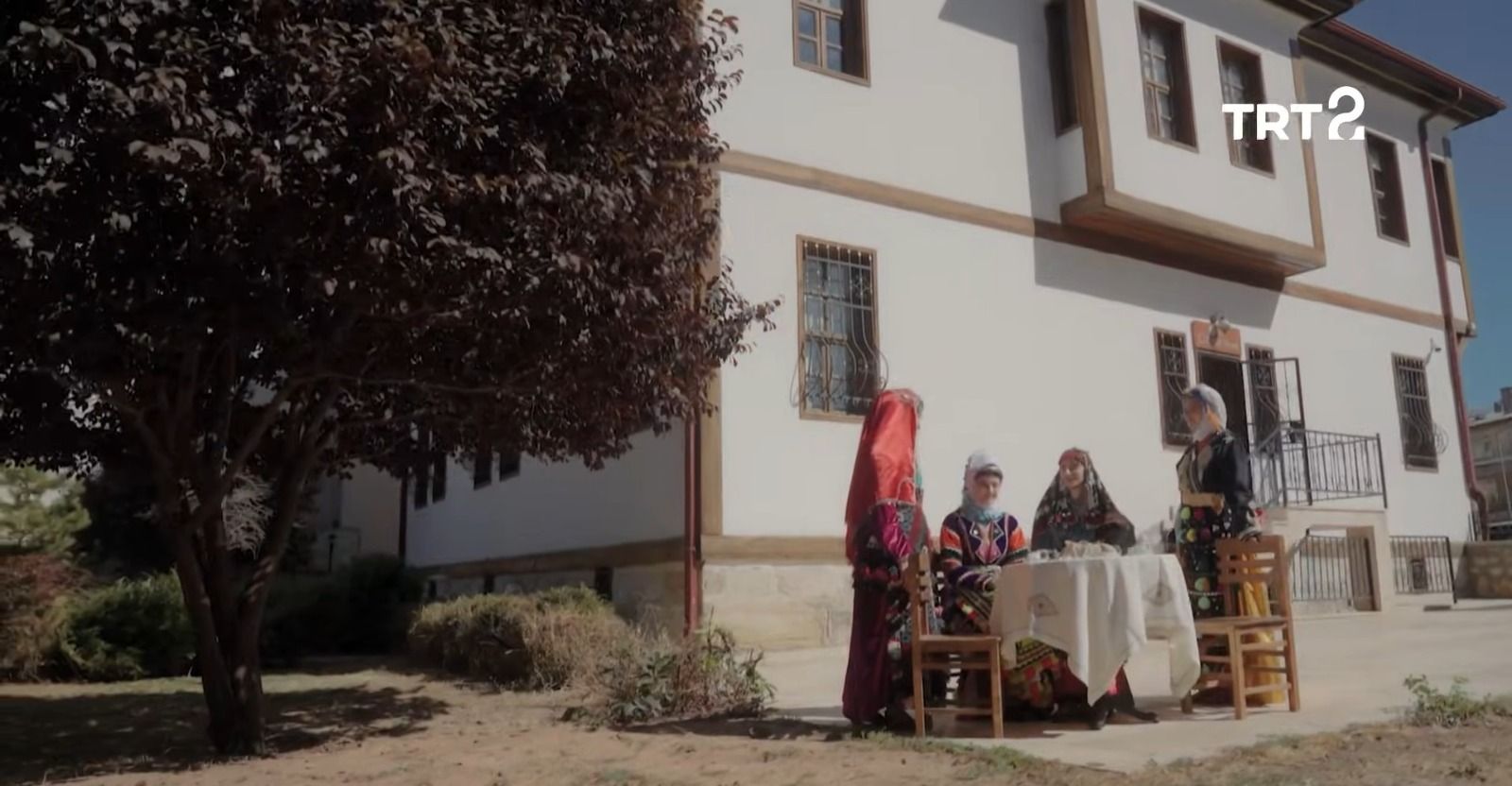 Bir binanın önünde geleneksel kıyafetler giyen üç kişi, masa ve sandalyelerin yanında duruyor.