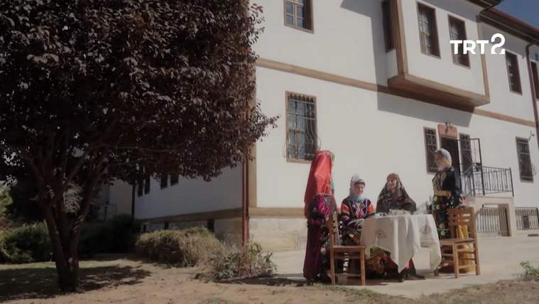 Bir binanın önünde geleneksel kıyafetler giyen üç kişi, masa ve sandalyelerin yanında duruyor.