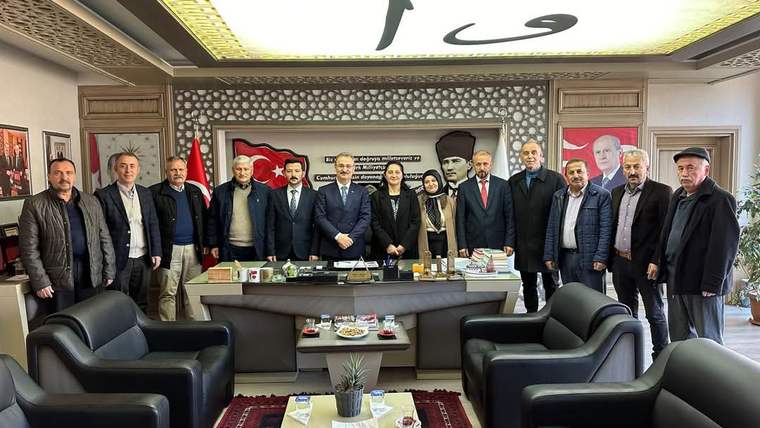 Bir ofiste masa arkasında duran bir grup kişi, Türk bayrağı ve duvarlardaki portrelerin önünde görülüyor.