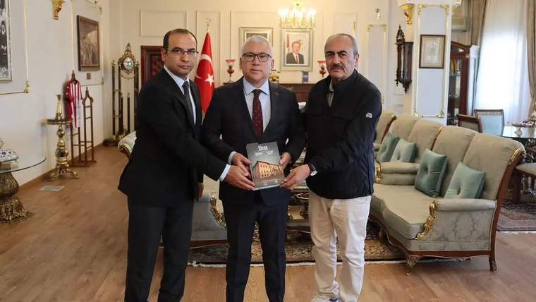 Üç kişi, bir kitap tutarak bir odada poz veriyor. Arka planda Türk bayrağı ve çeşitli mobilyalar var.