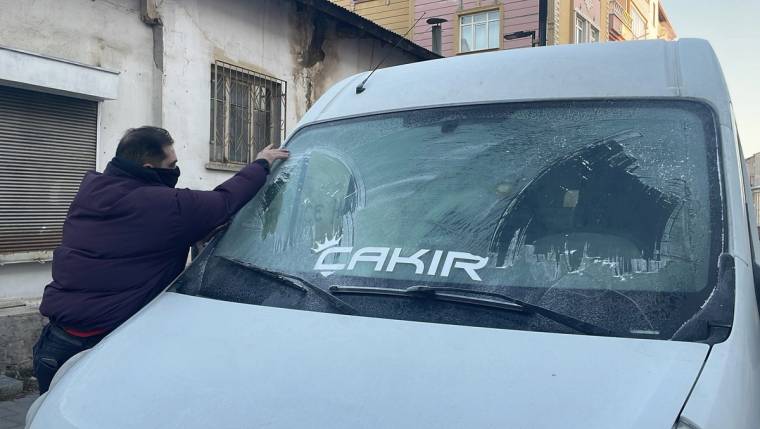Bir kişi, buz tutmuş minibüsün ön camını temizlerken görülüyor; camda "ÇAKIR" yazısı bulunuyor.