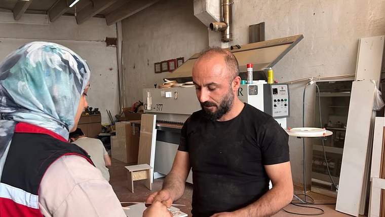 Başörtülü ve yelekli bir kadın, atölyede siyah tişörtlü sakallı bir erkekle tokalaşıyor.