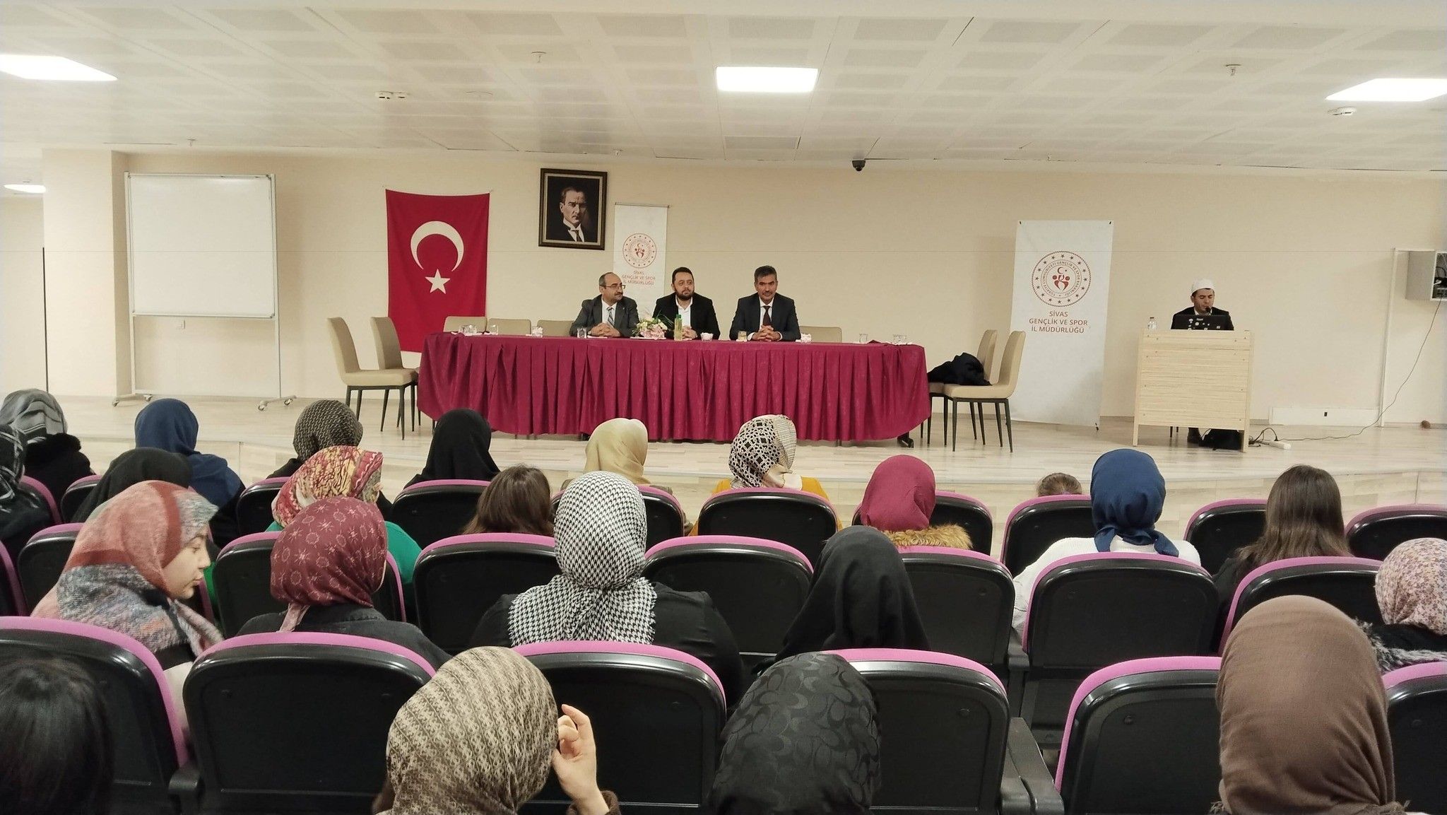 Bir grup kadın, sahnede oturan konuşmacıları dinliyor; arka planda Türk bayrağı ve Atatürk portresi var.