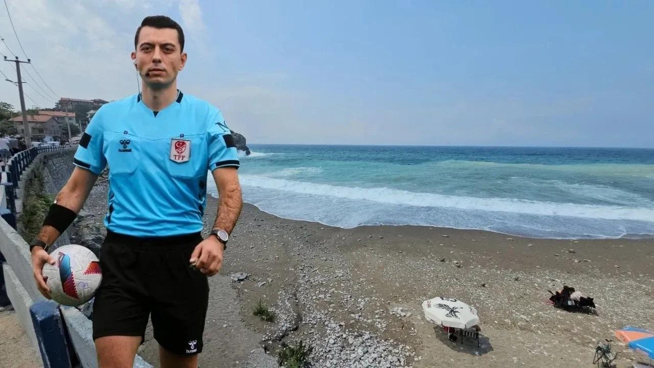 Mavi forması ve elinde futbol topuyla bir hakem, deniz kıyısındaki taşlı sahilde duruyor.