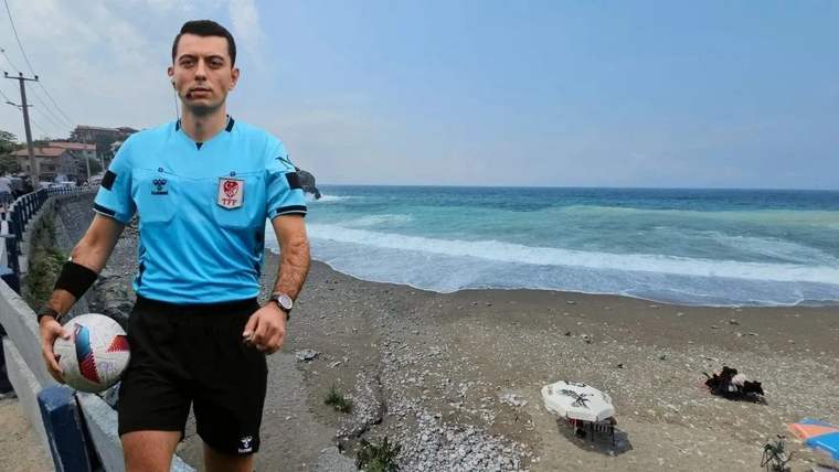 Mavi forması ve elinde futbol topuyla bir hakem, deniz kıyısındaki taşlı sahilde duruyor.