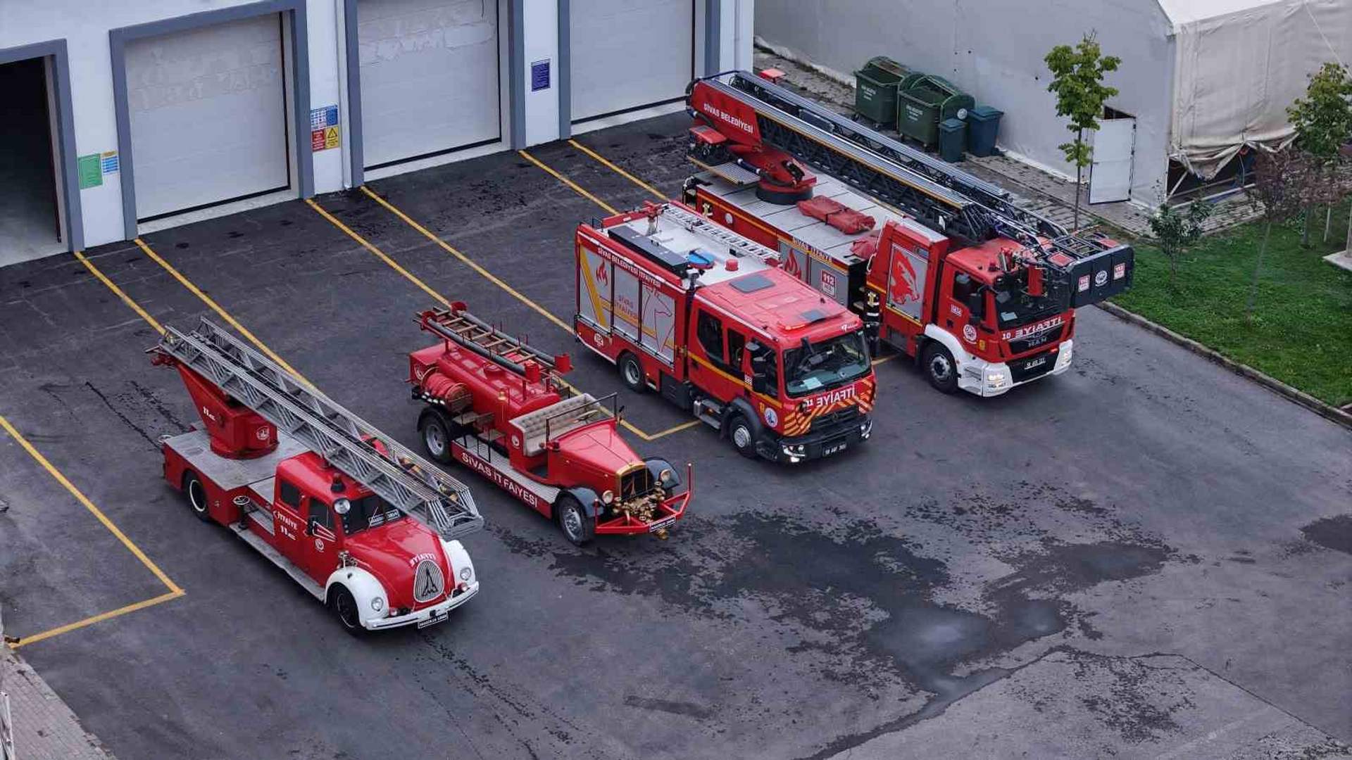 İtfaiye binasının önünde, farklı yıllara ait kırmızı itfaiye araçları ve merdivenli araçlar sıralanıyor.
