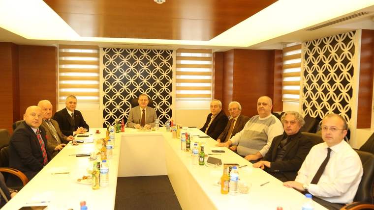 Toplantı masası çevresinde oturan bir grup kişi, önlerindeki su şişeleri ve dosyalarla birlikte görülüyor.