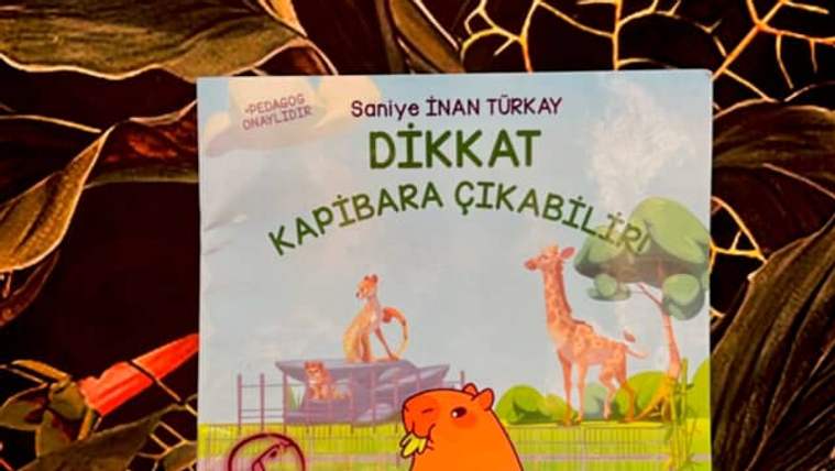 Kapakta turuncu bir kapibara, zürafa ve başka hayvan çizimleri bulunan çocuk kitabı, desenli zeminde duruyor.