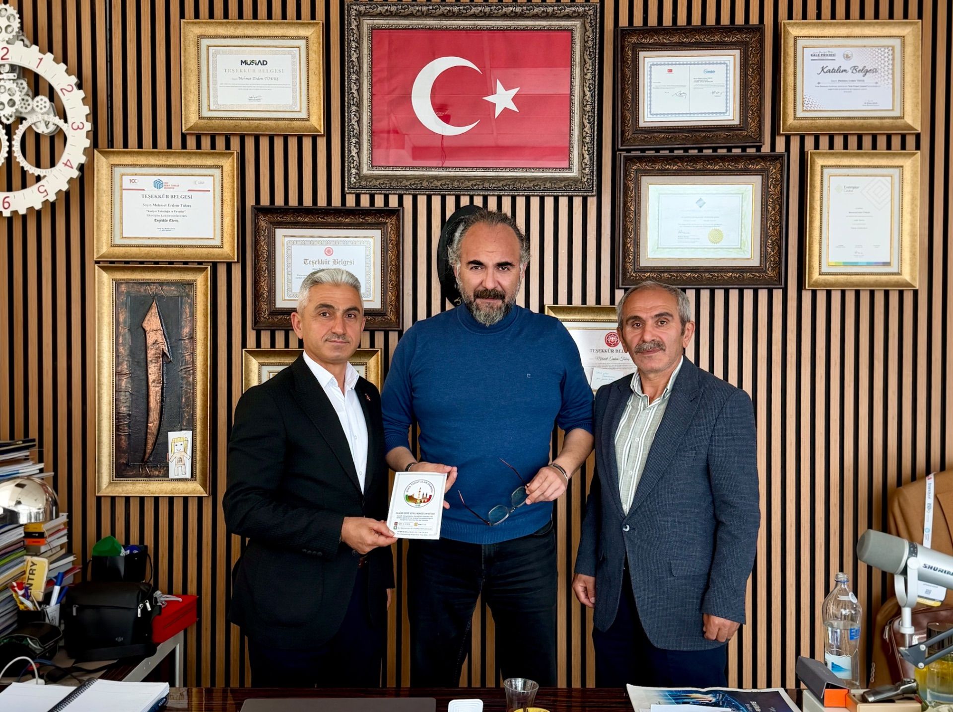 Üç erkek, duvarda asılı belgeler ve Türk bayrağının önünde duruyor; ortadaki kişi elinde kitapçık tutuyor.