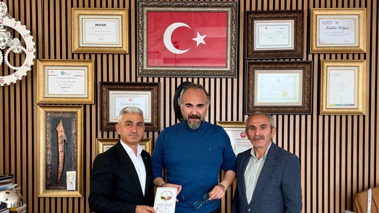 Üç erkek, duvarda asılı belgeler ve Türk bayrağının önünde duruyor; ortadaki kişi elinde kitapçık tutuyor.