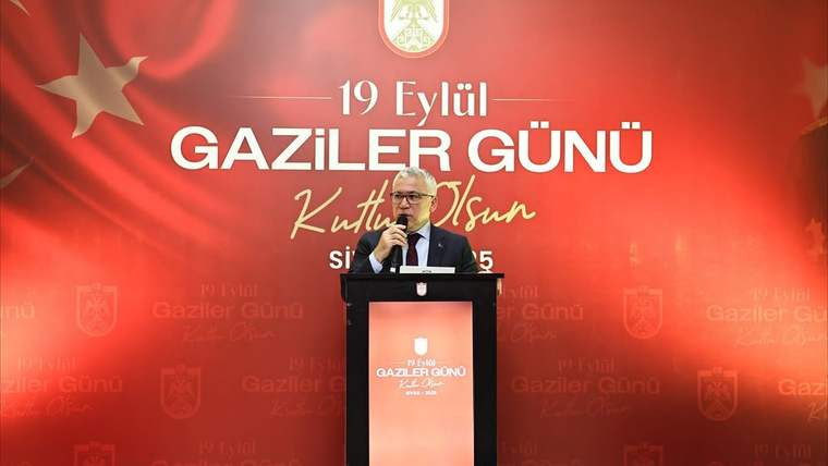 Kırmızı fonda 19 Eylül Gaziler Günü yazısı önünde, kürsüde mikrofonla konuşan bir kişi görülüyor.