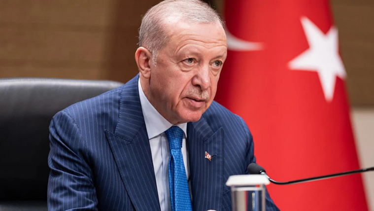 Mavi çizgili takım elbise giymiş bir kişi, masanın arkasında konuşuyor; arkasında Türk bayrağı var.