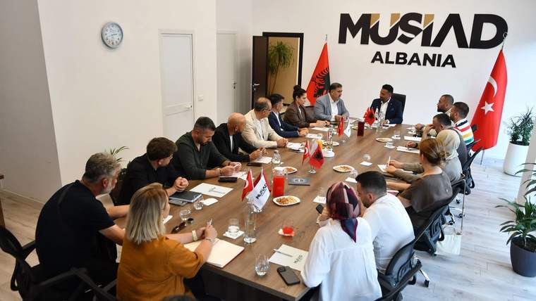 Bir toplantı odasında, uzun bir masanın etrafında oturan katılımcılar, not alıyor ve tartışıyor.