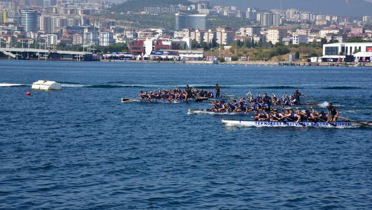 Denizde kürek çeken sporcuların bulunduğu uzun tekneler, kıyıdaki binalar ve dağlar önünde ilerliyor.