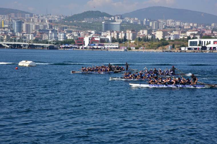 Denizde kürek çeken sporcuların bulunduğu uzun tekneler, kıyıdaki binalar ve dağlar önünde ilerliyor.