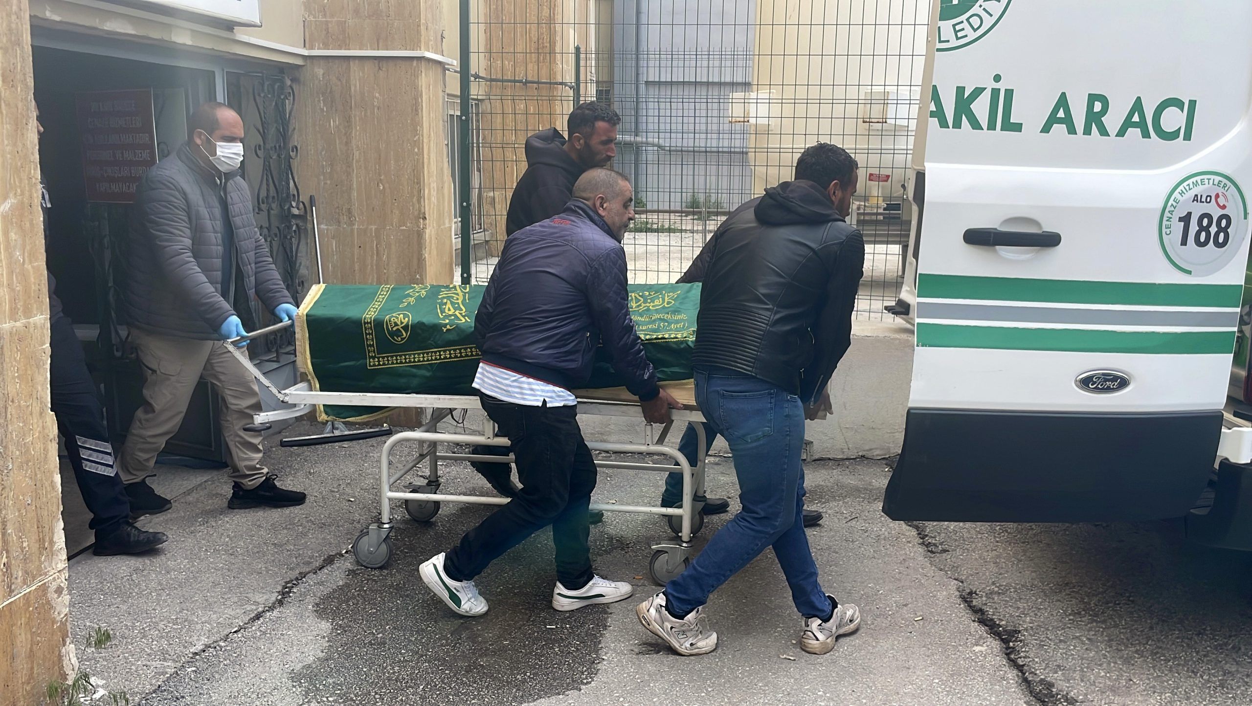 Bir grup insan, yeşil örtüyle kaplı bir tabutu hastane aracına taşıyor.
