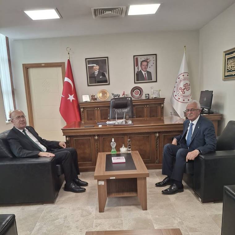 Makam odasında iki takım elbiseli kişi karşılıklı koltuklarda oturuyor; arkada Türk bayrağı ve kurum bayrağı bulunuyor.